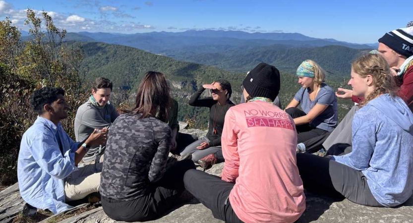 A group of people sit on a rock overlook. Below them is a vast green mountainous area. 